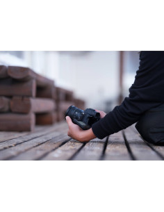 A photographer kneels on wooden planks, holding a camera and lens, in a blurred outdoor s, captured with Sony FE 16mm f/1.8 G A photographer kneels on wooden planks, holding a camera and lens, in a blurred outdoor s, captured with Sony FE 16mm f/1.8 G