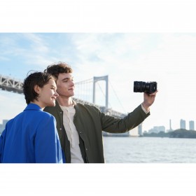 Un couple souriant prend un selfie avec un pont en arrière-plan sous un ciel bleu, capture au Sony FE 20-70mm f/4 G